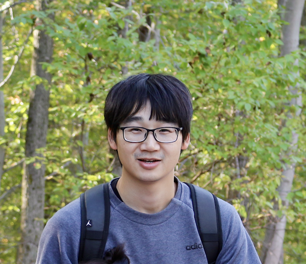 photo of young man with dark hair and glasses standing in from of green trees